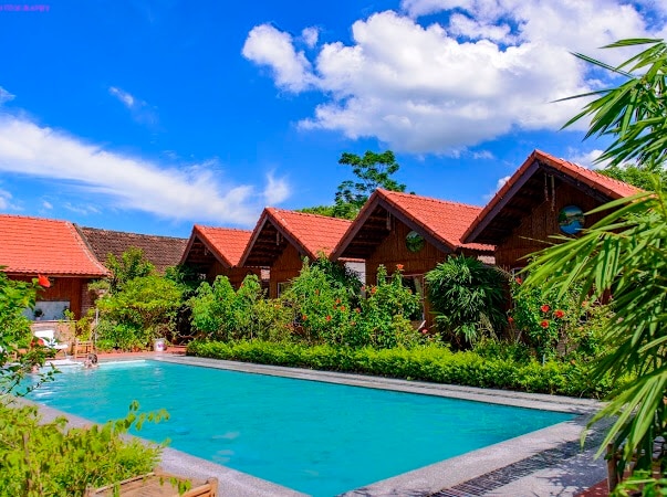 A communal swimming pool is surrounded by lush greenery and blooming plants, with wooden cottages featuring slanted roofs visible in the background. The clear blue water reflects the sky, and the inviting pool area is enhanced by the natural setting.