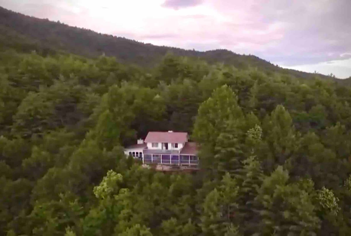 An aerial view captures a spacious cabin nestled among lush greenery, surrounded by trees and rolling mountains. The structure features a red roof and multiple balconies, emphasizing its secluded setting within the natural landscape.