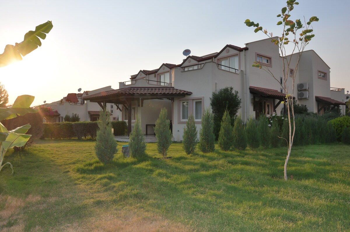 A two-story villa is depicted, featuring a well-maintained lawn and lush greenery surrounding the property. The structure showcases a terrace area with a shaded entrance, set against a backdrop of a soft, glowing sunset.