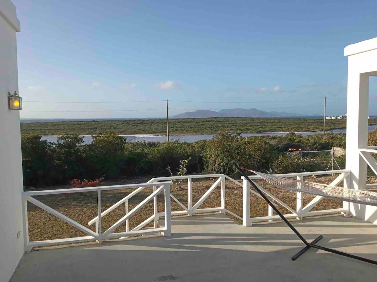 A spacious outdoor terrace offers serene views of the surrounding landscape, featuring greenery and distant mountains under a clear blue sky. A hammock is set up, inviting relaxation while enjoying the tropical scenery.