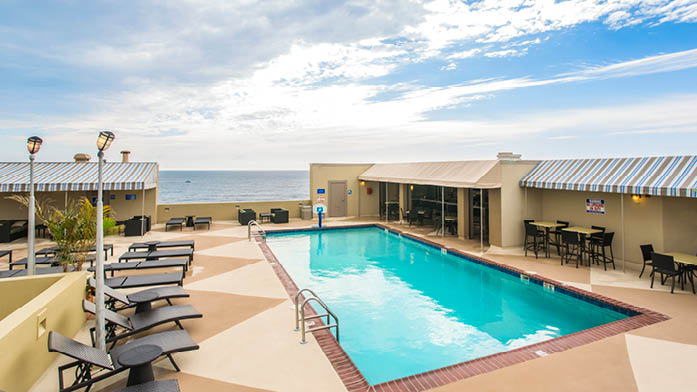An outdoor pool is surrounded by a patio featuring lounge chairs and seating areas. The ocean can be seen in the background under a partly cloudy sky. A poolside bar area is located to the left, providing a space for relaxation and refreshments.