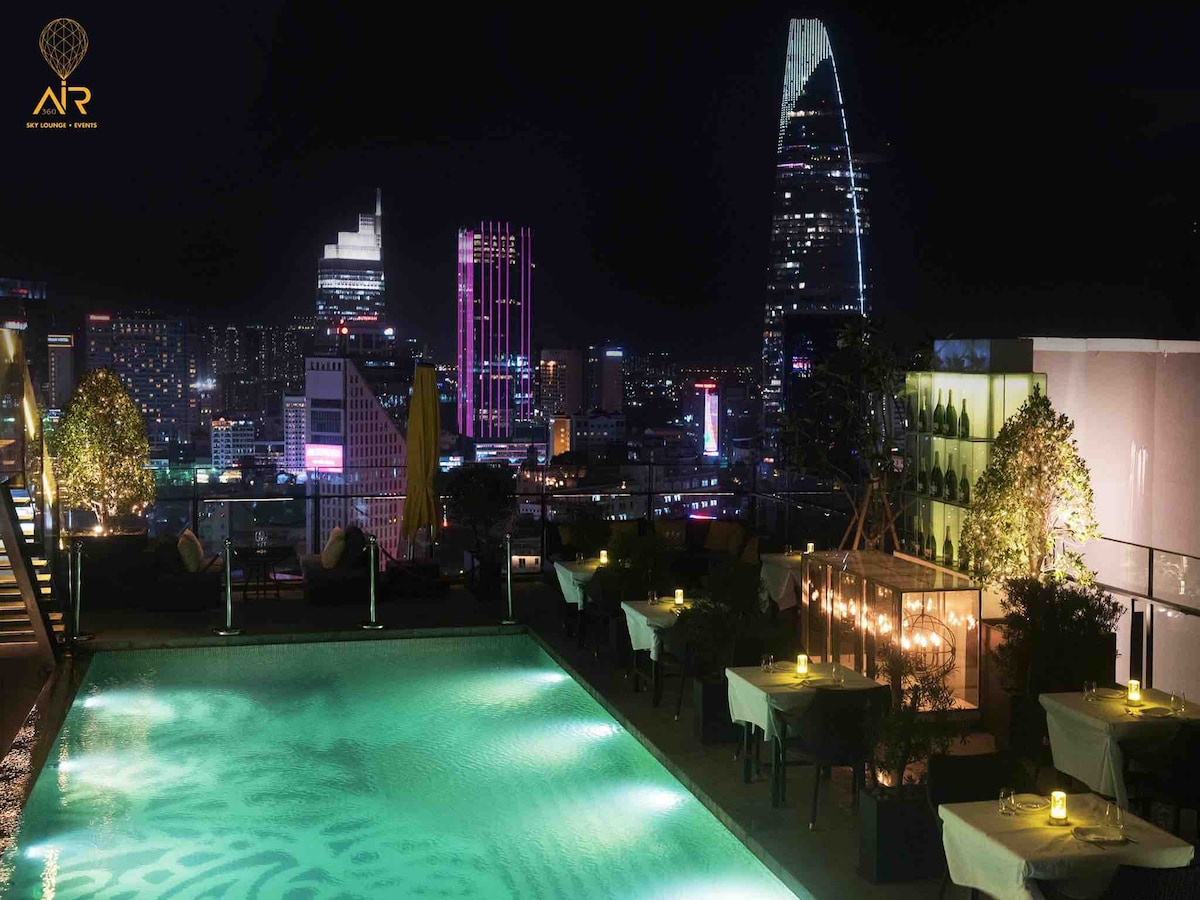 The rooftop pool glimmers under the night sky, surrounded by comfortable seating and small tables lit by soft candlelight. The city skyline is visible in the background, featuring a blend of colorful lights from nearby buildings.