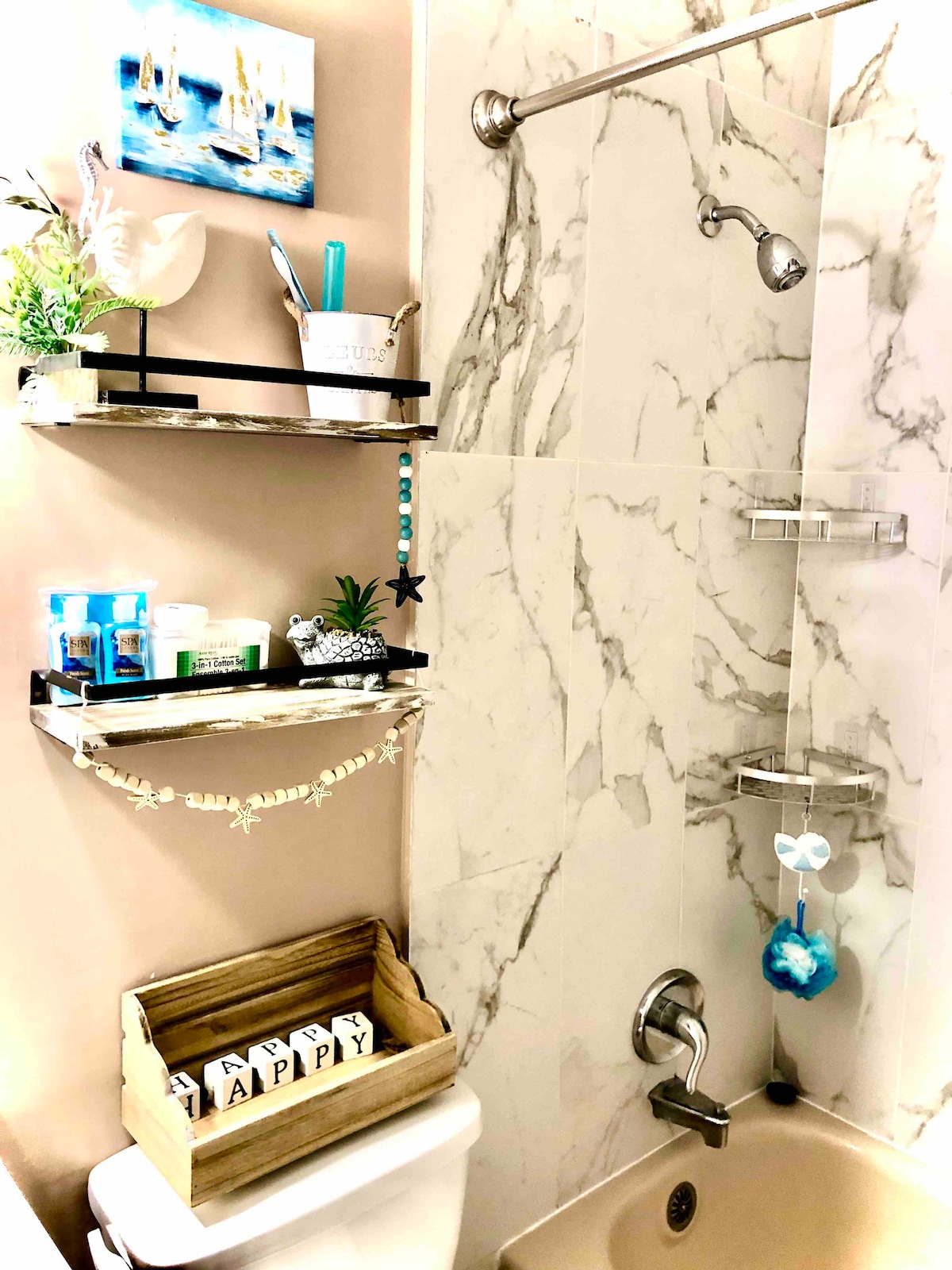 A bathroom is presented with a modern marble wall and a showerhead mounted above. Shelves are equipped with toiletries and decorative items. A wooden crate holds blocks that spell 'HAPPY', contributing to a lighthearted atmosphere. Decorative plants enhance the space.