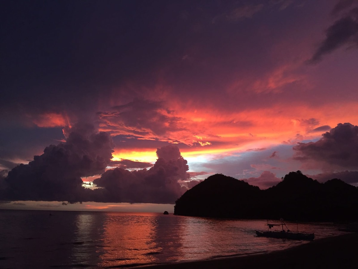 A vibrant sunset illuminates the sky with hues of orange, pink, and purple, reflecting on the calm waters. Silhouettes of distant mountains and clouds create a dramatic backdrop, enhancing the tranquil beach panorama.