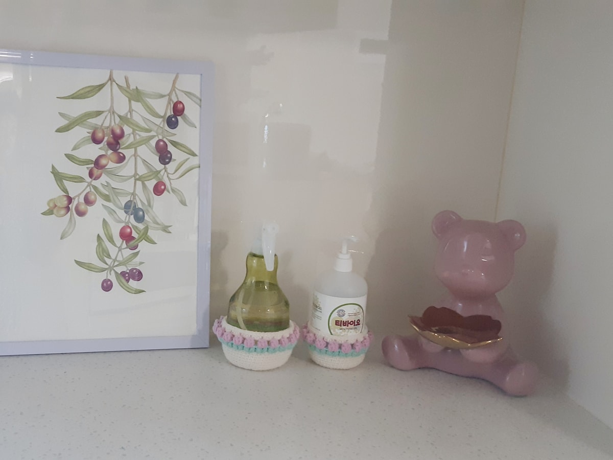 A countertop display features a decorative framed illustration of olives beside two small bottles, one for oil and the other for soap. A pink ceramic bear figurine holds a shallow dish, adding a touch of whimsy to the peaceful arrangement.