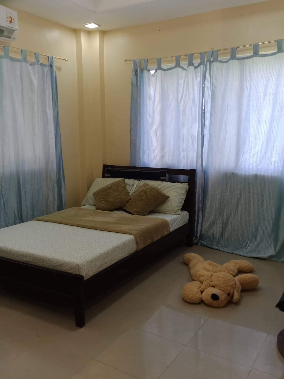 A bright bedroom features a single bed with decorative pillows, set against a wall of soft yellow. Light blue curtains filter natural light from a window, creating a serene atmosphere. A plush teddy bear is positioned on the floor, adding a playful touch to the space.