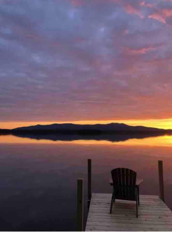 A serene scene features a wooden dock extending over calm waters, with a single black Adirondack chair facing the vibrant sunrise. Soft clouds reflect shades of orange and purple in the sky above the distant mountains, creating a peaceful atmosphere.