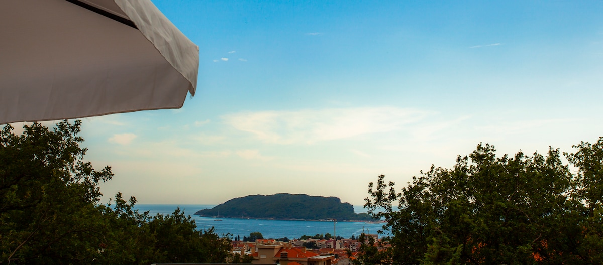 A panoramic view of the blue sea and a distant island is framed by vibrant greenery. The sky is clear, showcasing shades of blue, while the horizon merges the sea and sky. Quiet residential areas are visible in the foreground.