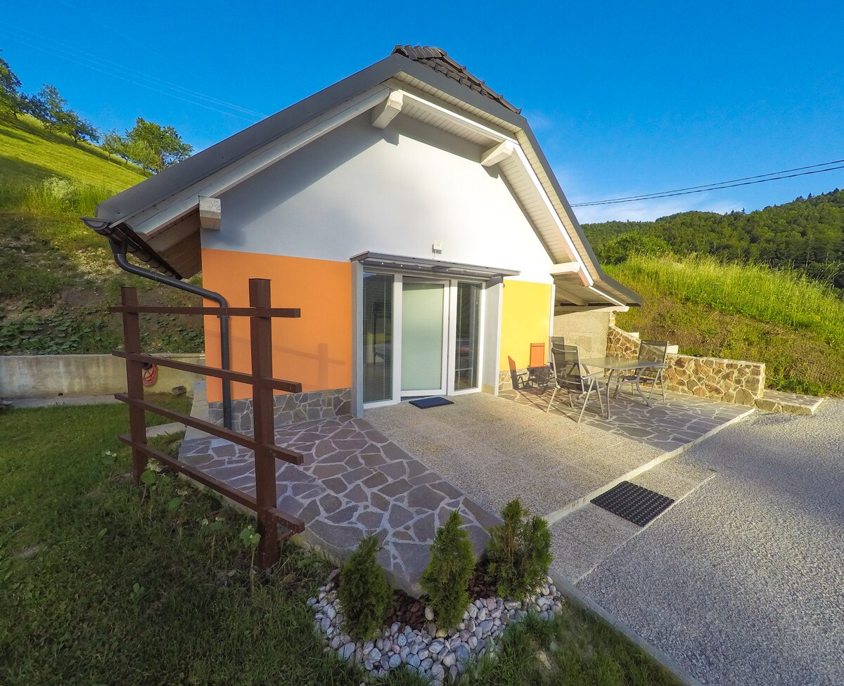 A modern cabin exterior is showcased, featuring a combination of orange and grey walls. Outdoor seating is arranged on a stone patio, accompanied by a small table and two chairs. Lush greenery surrounds the area, enhancing the peaceful environment.