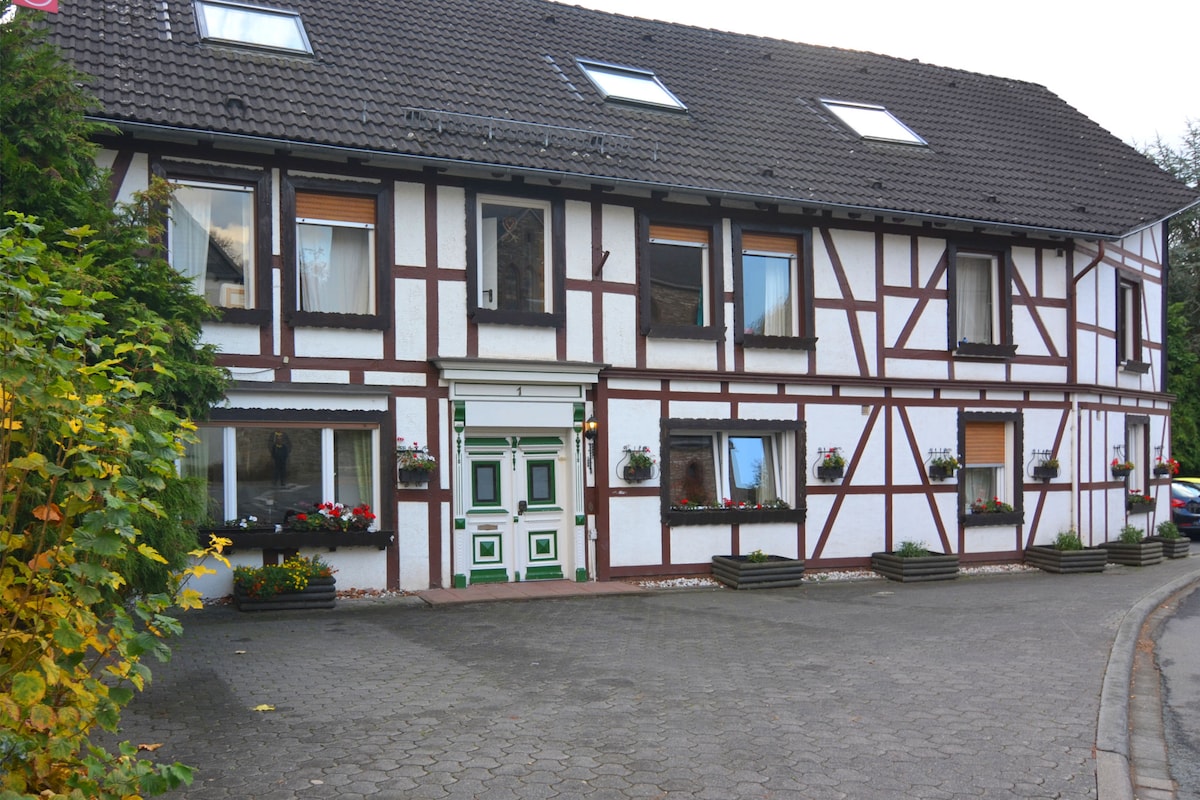 A charming two-story holiday home features a half-timbered facade with white walls and dark wood accents. Flower boxes adorn the windows, and a welcoming green and white door is centered at the entrance. The landscaped area includes greenery along the driveway.