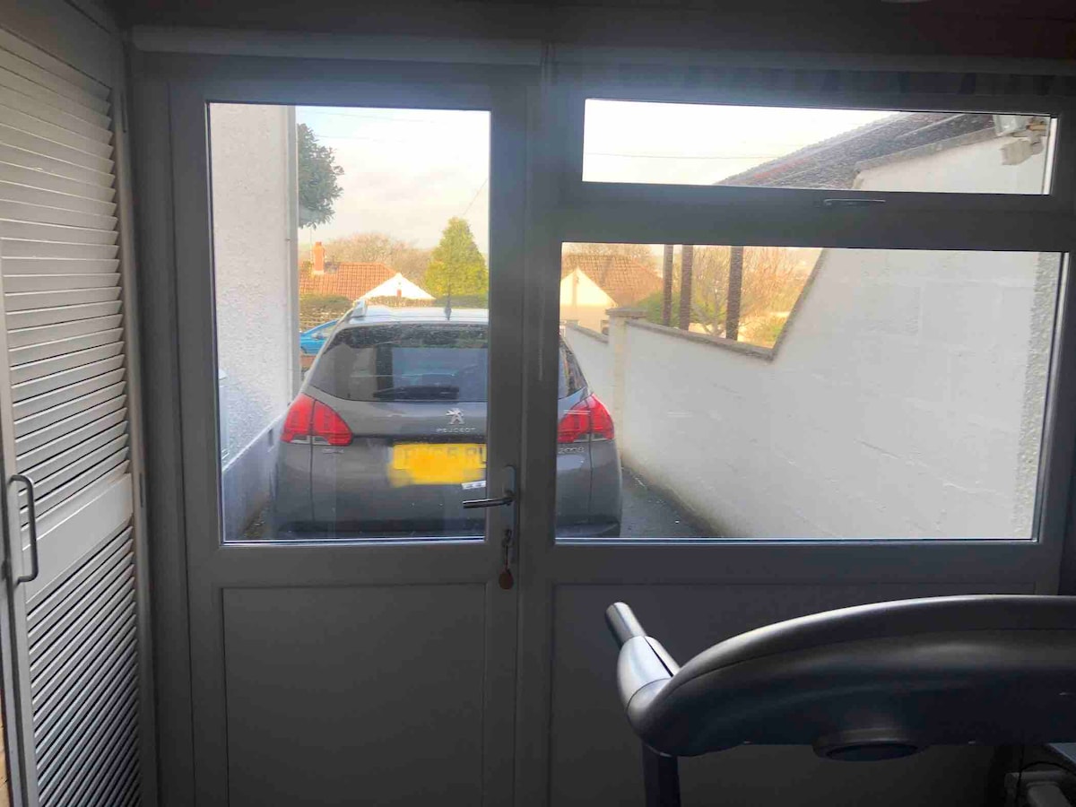 A covered garden room features large windows, providing a view of the driveway with a parked car. The interior includes an exercise bike, and natural light filters in through the glass, illuminating the space.