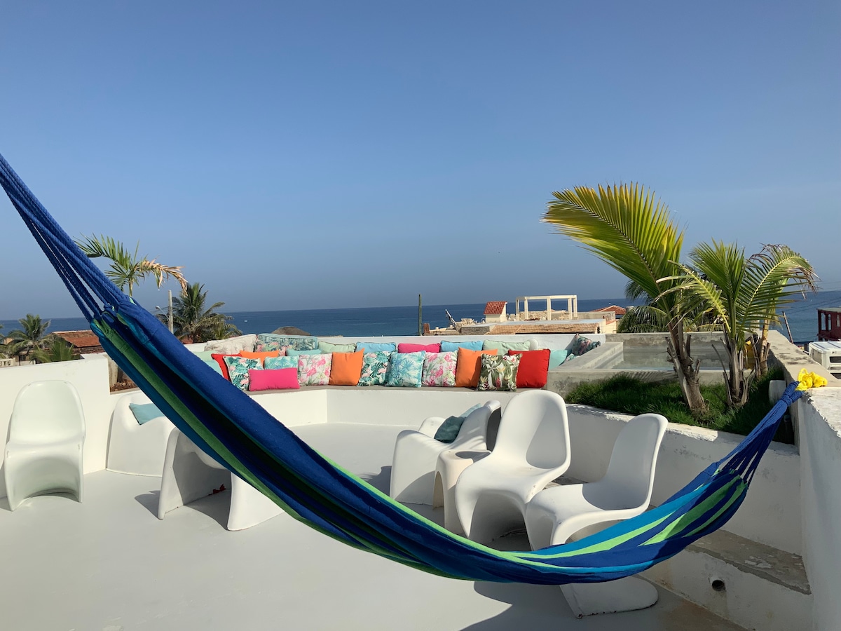 A spacious terrace features a colorful hammock and white seating, overlooking the ocean. Bright cushions add splashes of color against the serene backdrop. The small pool can be seen in the distance, framed by lush green palm trees and coastal views.
