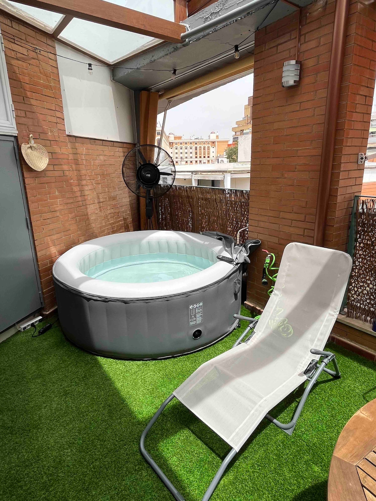 A private terrace features an inflatable jacuzzi surrounded by green artificial grass. A sun lounger is positioned nearby, and a ceiling fan is visible, providing a cooling breeze. The backdrop highlights the urban skyline, with sunlight illuminating the area.