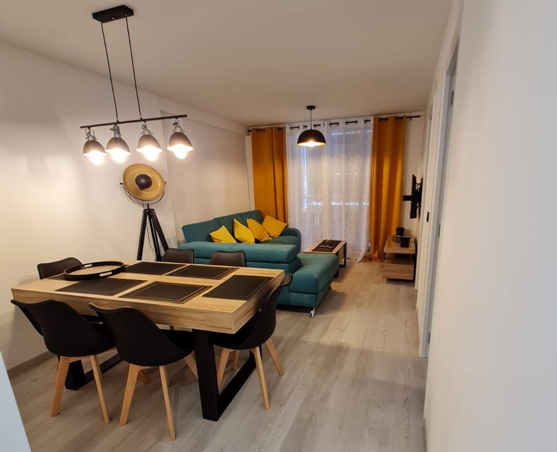 A modern living area features a spacious dining table paired with black chairs. A teal sofa is adorned with yellow throw pillows, providing a pop of color against the white walls. Illuminated by pendant lighting, the area is designed for relaxation and casual dining.