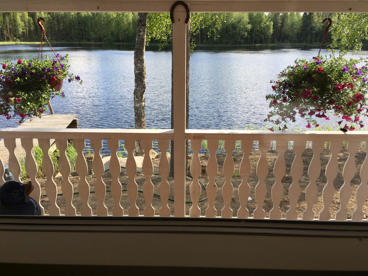 A view from a porch is presented, showcasing a serene lake bordered by trees. Hanging flower pots filled with vibrant blooms add color to the scene. A wooden deck extends toward the water, enhancing the connection between the indoor space and the natural surroundings.