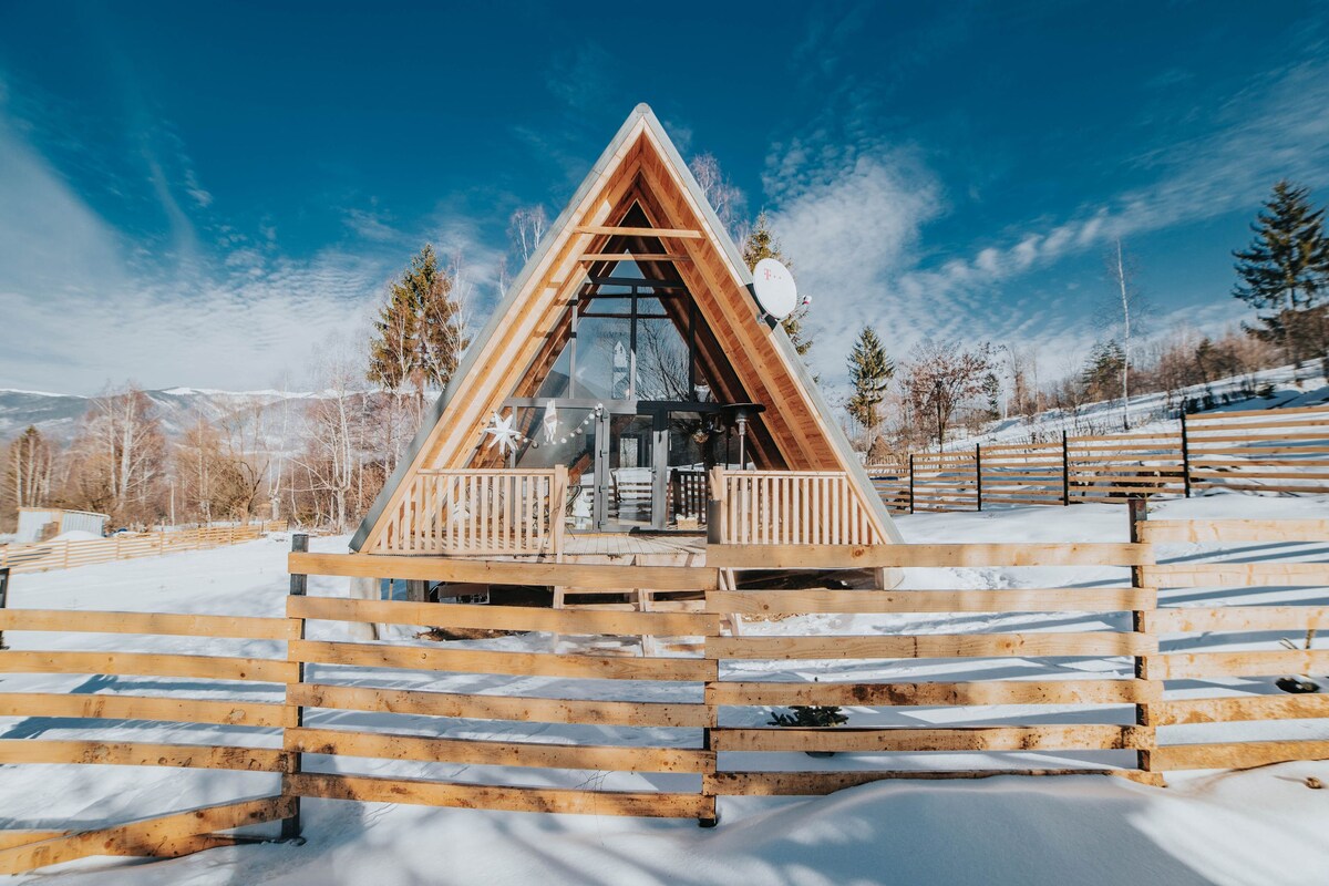 An A-frame cabin is showcased amidst a winter landscape, featuring a wooden fence surrounding the property. The cabin is elevated, with large windows offering views of the surroundings, and snow blankets the ground, enhancing the serene outdoor setting.