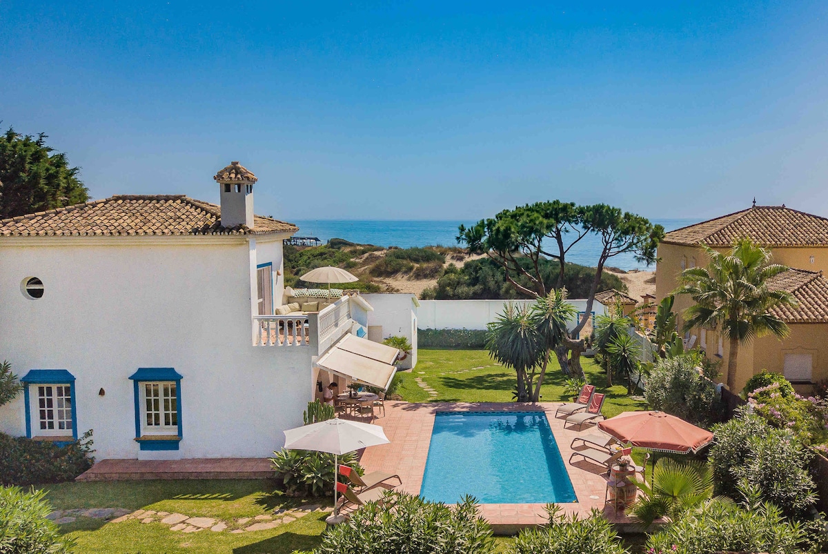 A charming villa is situated near the coast, featuring a clear blue swimming pool surrounded by green foliage. Several lounge chairs and umbrellas are arranged around the pool. In the background, the ocean is visible alongside sandy shores and residential structures.