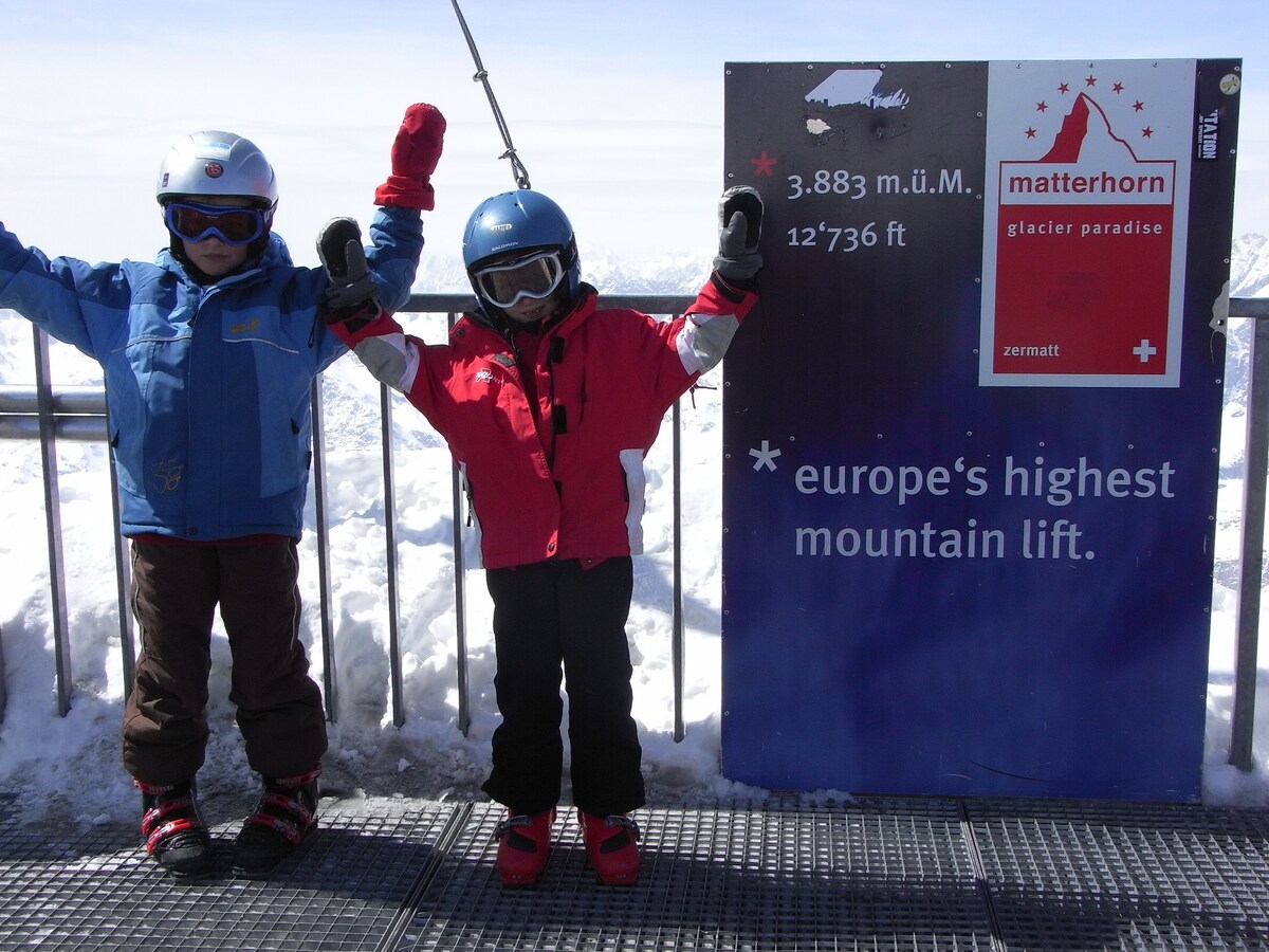 Two children stand joyfully at a mountain viewpoint, wearing helmets and snow gear. A sign beside them indicates the altitude of 3,883 m and displays information about the Matterhorn Glacier Paradise. Snow-covered terrain stretches into the distance, highlighting the alpine setting.
