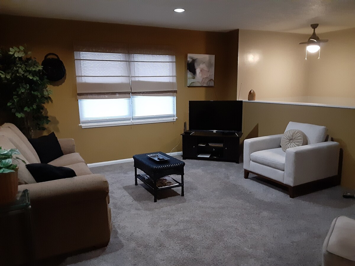 The living area features a neutral color palette with comfortable seating arrangements, including a beige sofa and a white accent chair. A coffee table sits centrally, and a television is positioned against the wall. Natural light enters through two windows adorned with shades.
