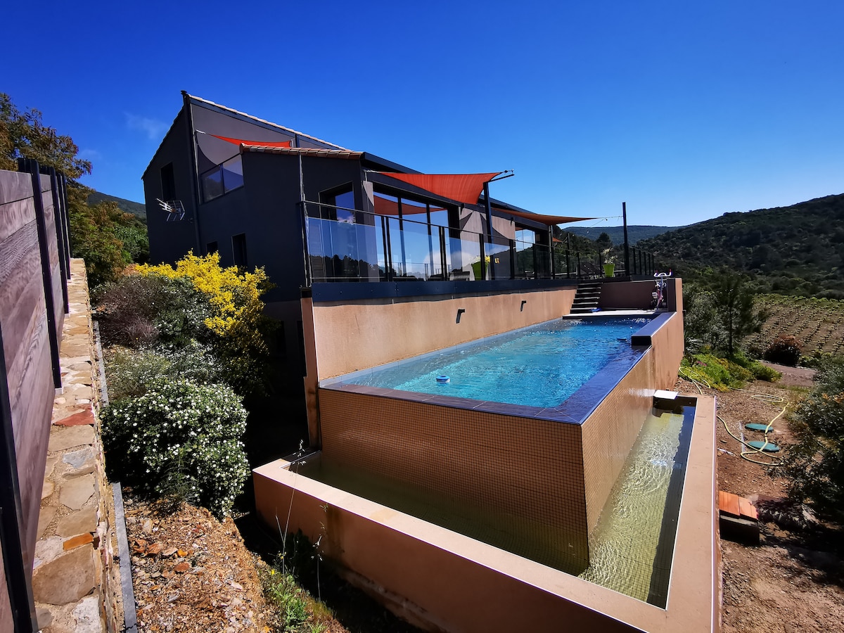 The modern house is situated on a hillside, overlooking vineyards. A private infinity pool extends from the structure, surrounded by a glass railing. Sunshades add color against the clear blue sky, emphasizing the tranquil outdoor space.