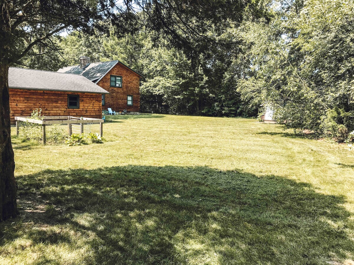 A spacious grassy area is surrounded by trees, providing a sense of privacy. In the background, a wooden house and a smaller structure are partially visible, blending harmoniously with the natural landscape.