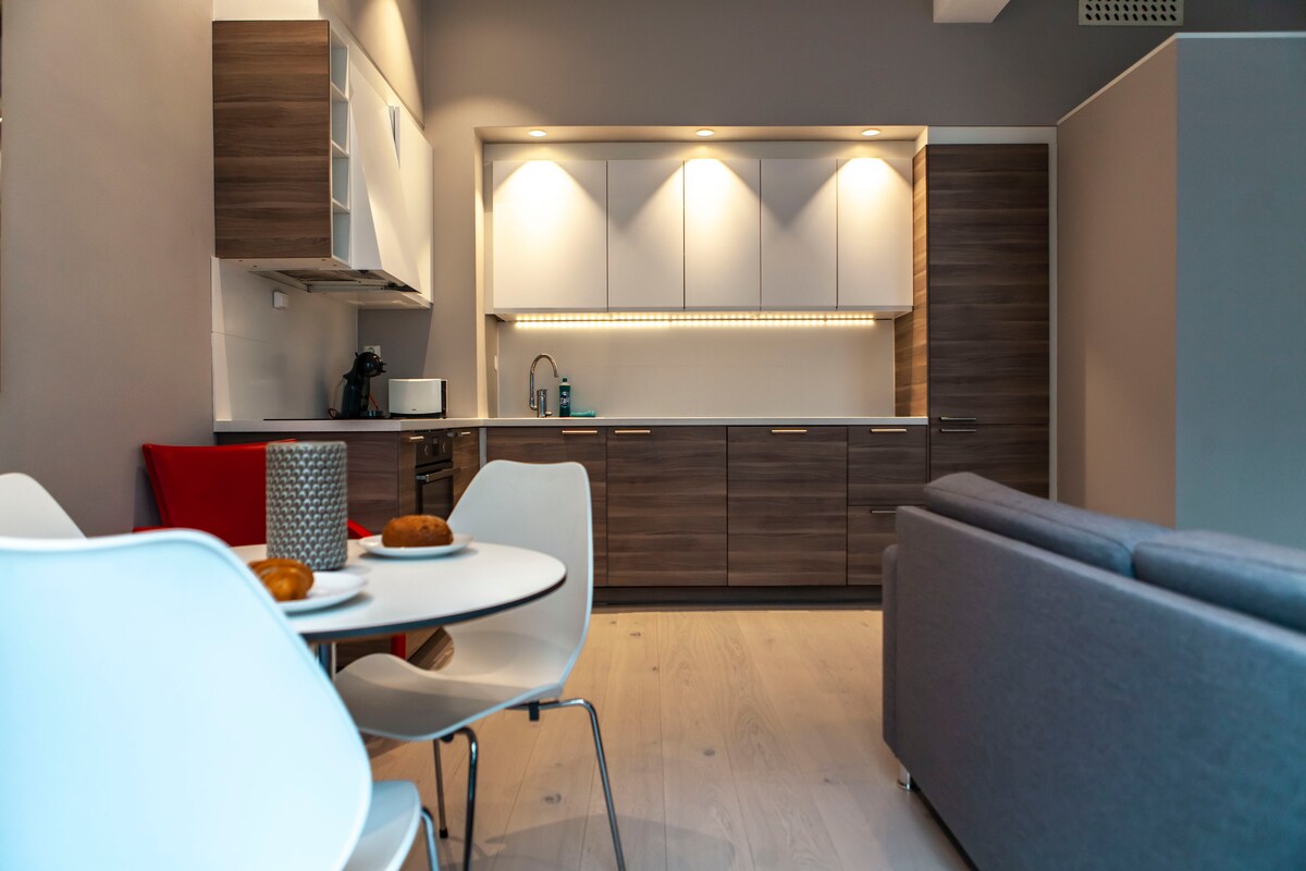 An open plan kitchen and living area features modern cabinetry and countertop. A round dining table with four white chairs is set with plates and pastries. The sofa in the living area is positioned comfortably, enhancing the spacious ambiance.