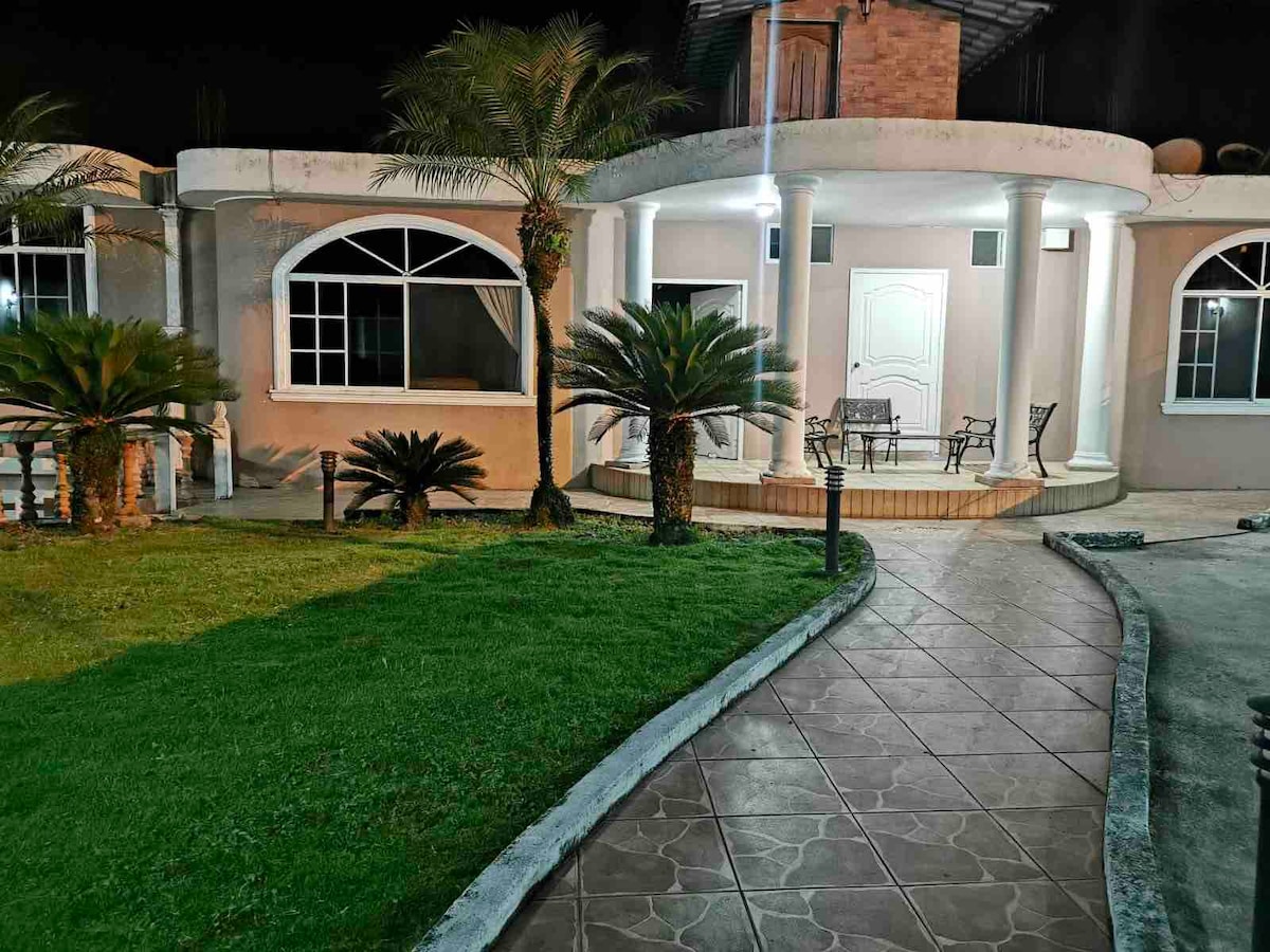The exterior of the house is illuminated at night, featuring a covered porch with seating. Lush palm trees are positioned around a well-maintained lawn, and a winding pathway leads toward the entrance. Large windows allow for natural light, creating an inviting ambiance.