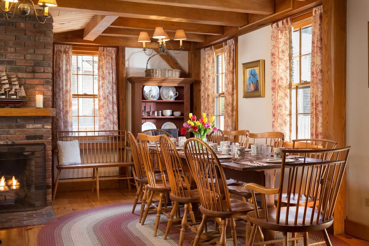 A welcoming dining area features a long wooden table surrounded by several chairs. Natural light enters through the windows, which are adorned with floral curtains. A cozy fireplace is visible, adding warmth to the space, and a bouquet of flowers decorates the table.