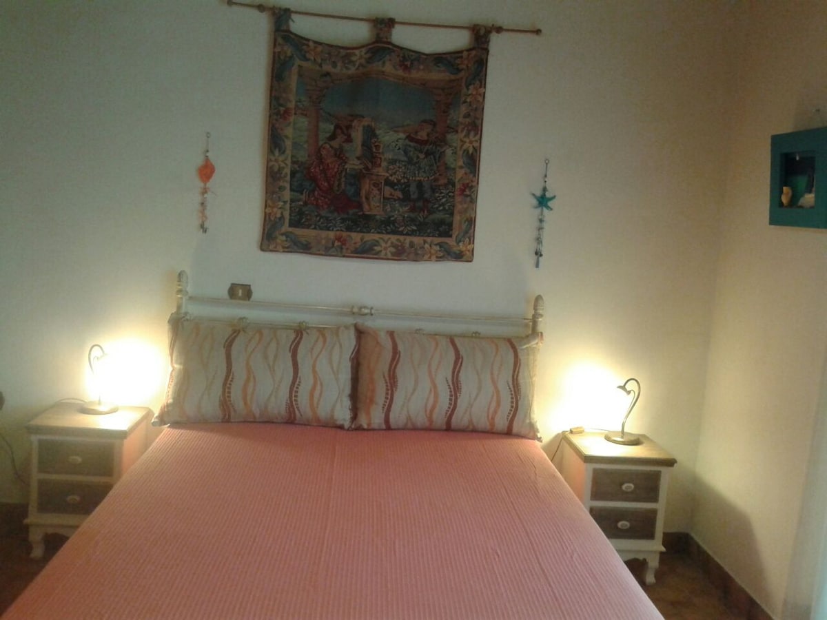 The master bedroom features a double bed with a coral-patterned blanket, flanked by two bedside tables with lamps. A decorative wall hanging adds character, while soft lighting creates a gentle ambiance.
