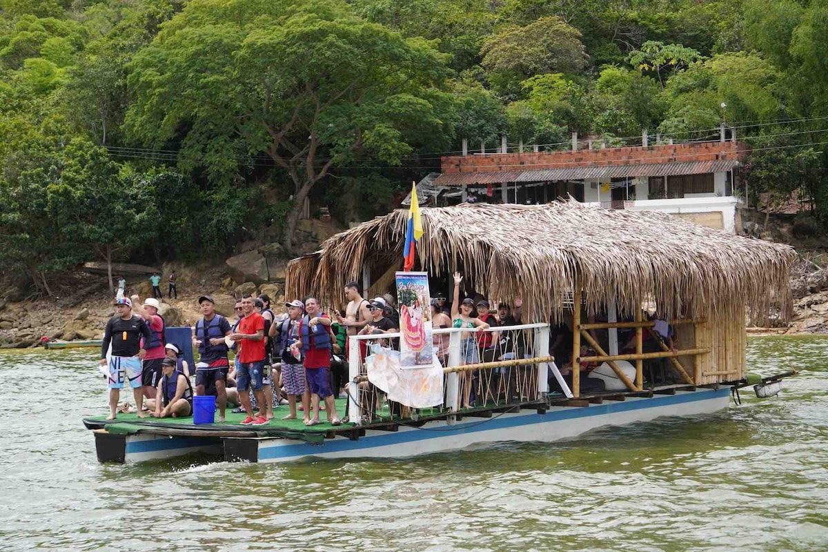 A spacious boat, topped with a thatched roof, is filled with people enjoying a day on the water. The vessel is anchored near lush green trees, with buildings visible on the shore in the background.