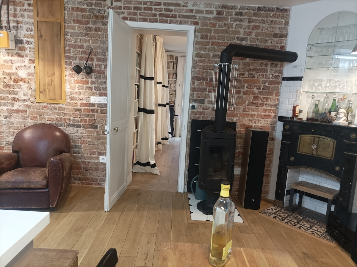 The living area features exposed brick walls and hardwood flooring. A rustic armchair is positioned nearby a wood stove, and a bottle is placed on a dining table. The image captures a view into a hallway lined with curtains, leading to additional spaces.