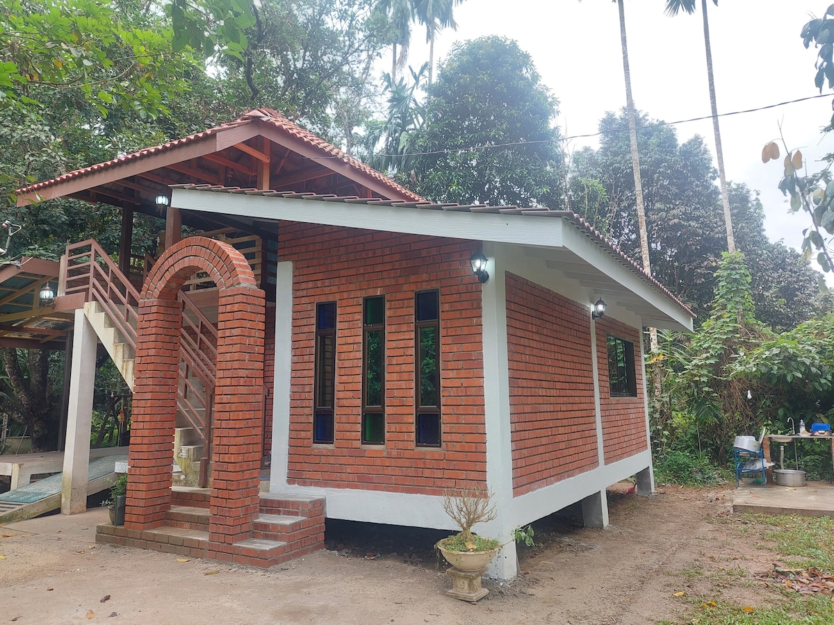 A cottage featuring a combination of red brick and white masonry stands amidst lush greenery. Large windows provide natural light, and a staircase leads to an upper outdoor area. The surrounding landscape includes trees and a pathway leading to additional outdoor facilities.