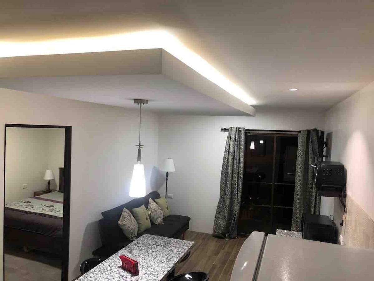 A well-lit living area features a cozy sofa adorned with decorative cushions, and a dining table with a stone-like surface. Stylish pendant lighting hangs above the table, and large windows are dressed in patterned curtains, allowing natural light to enter the space.