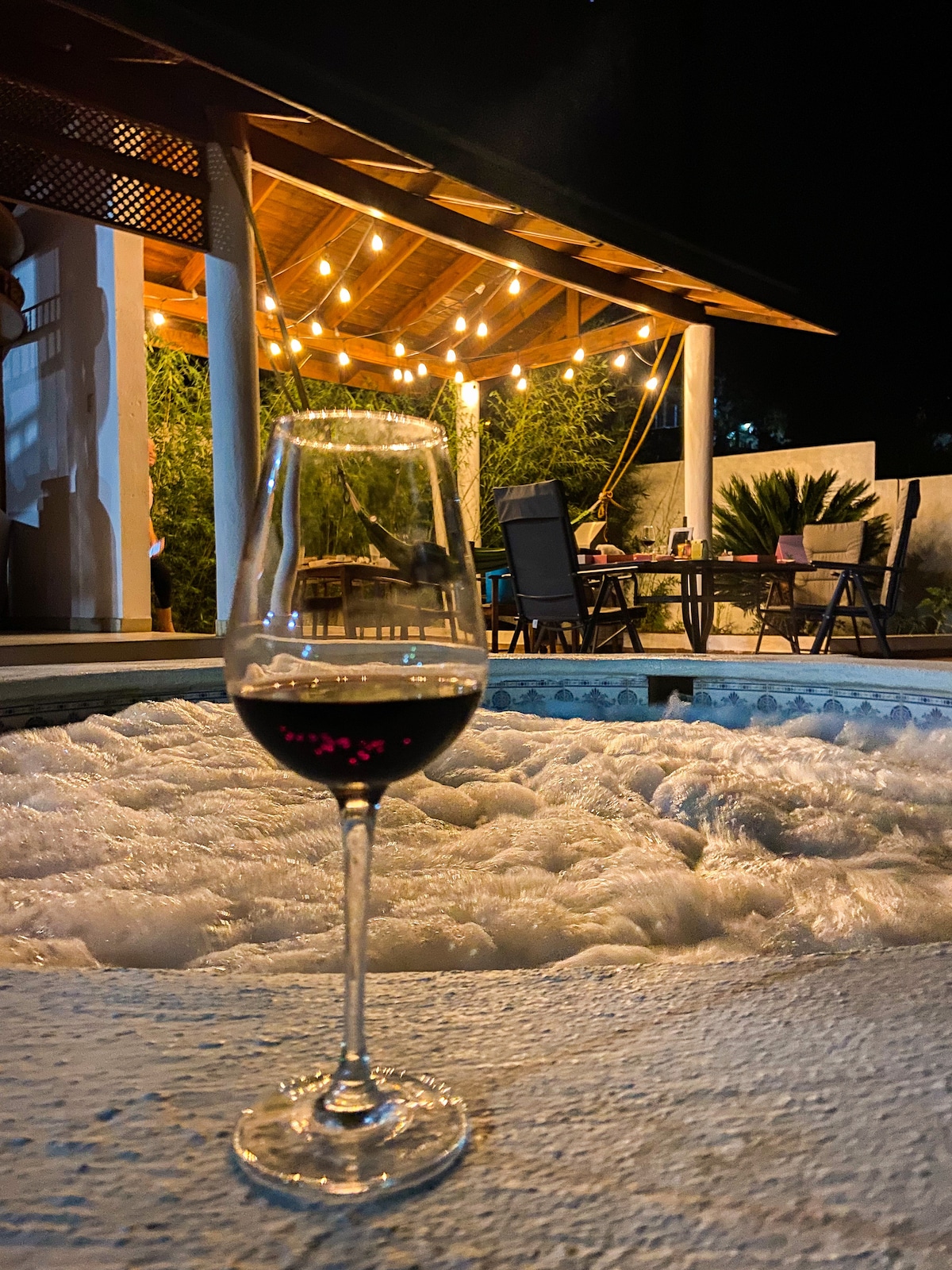 A glass of red wine is positioned in the foreground, showcasing its rich color against the backdrop of a softly bubbling jacuzzi. The illuminated terrace reveals a dining table and chairs under warm string lights, creating a welcoming outdoor space for relaxation.