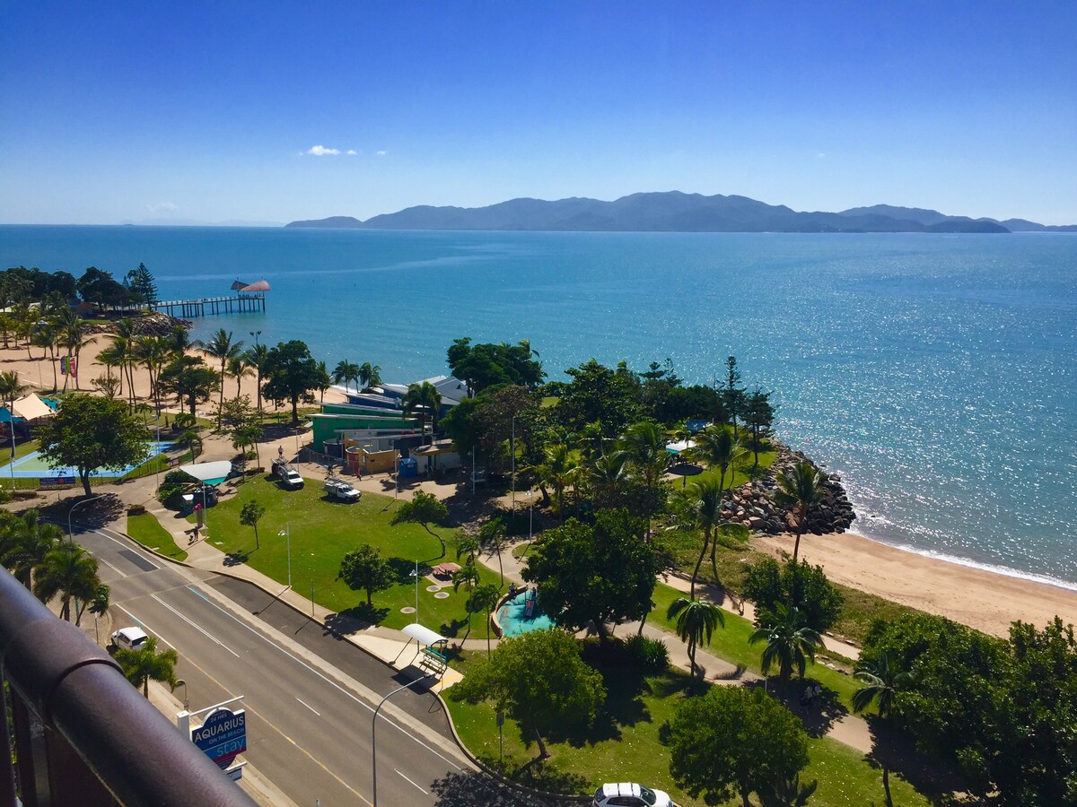 The image captures a panoramic view of the coastline featuring clear blue water and lush greenery. A sandy beach stretches along the shore, while an amusement park with pools is visible nearby. The distant mountains add depth to the serene landscape.