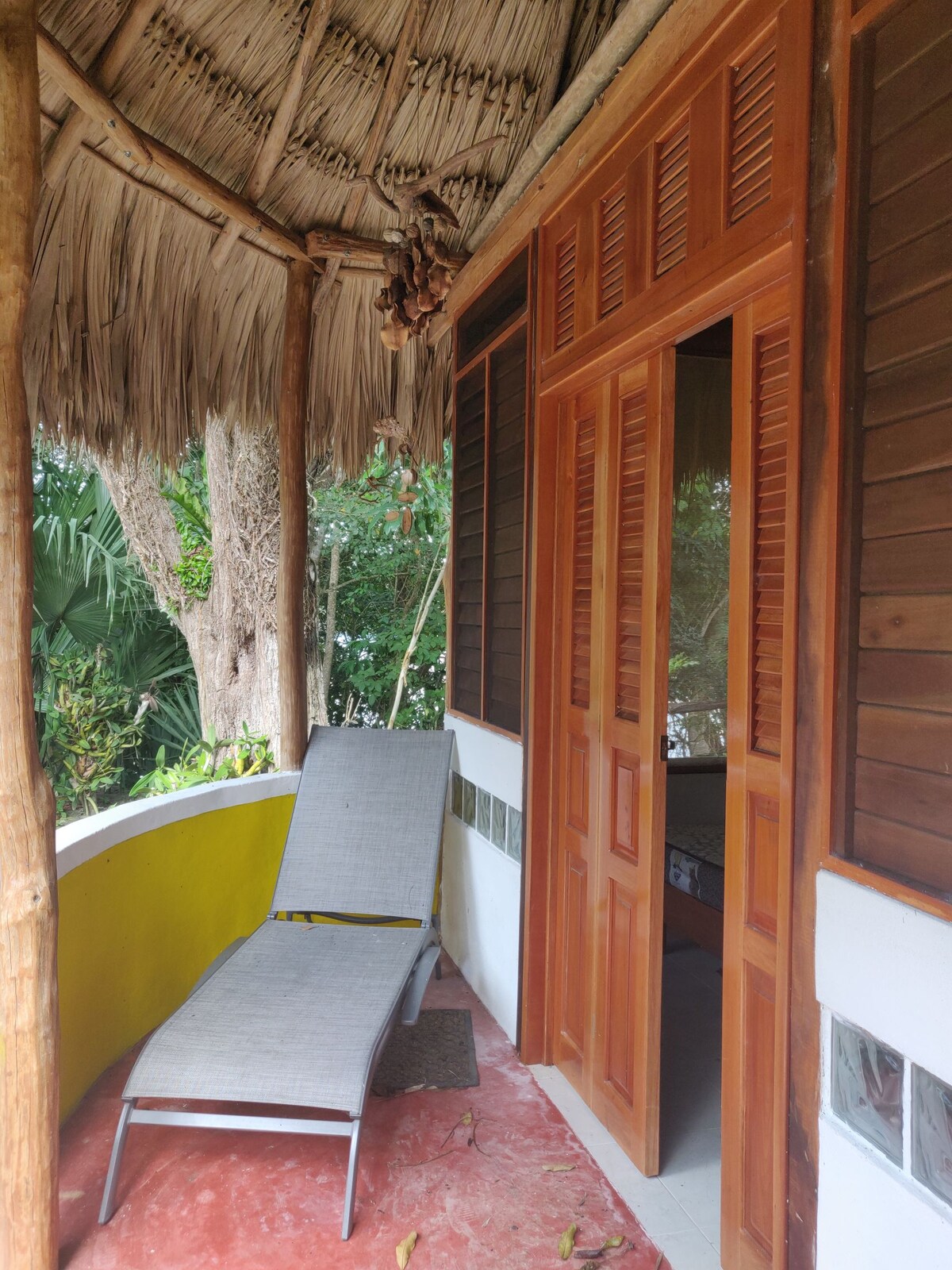 A shaded porch area features a single lounge chair made of grey fabric, positioned on a red-tiled surface. The entrance to a room is visible, flanked by wooden doors and surrounded by lush greenery, which provides a serene backdrop.