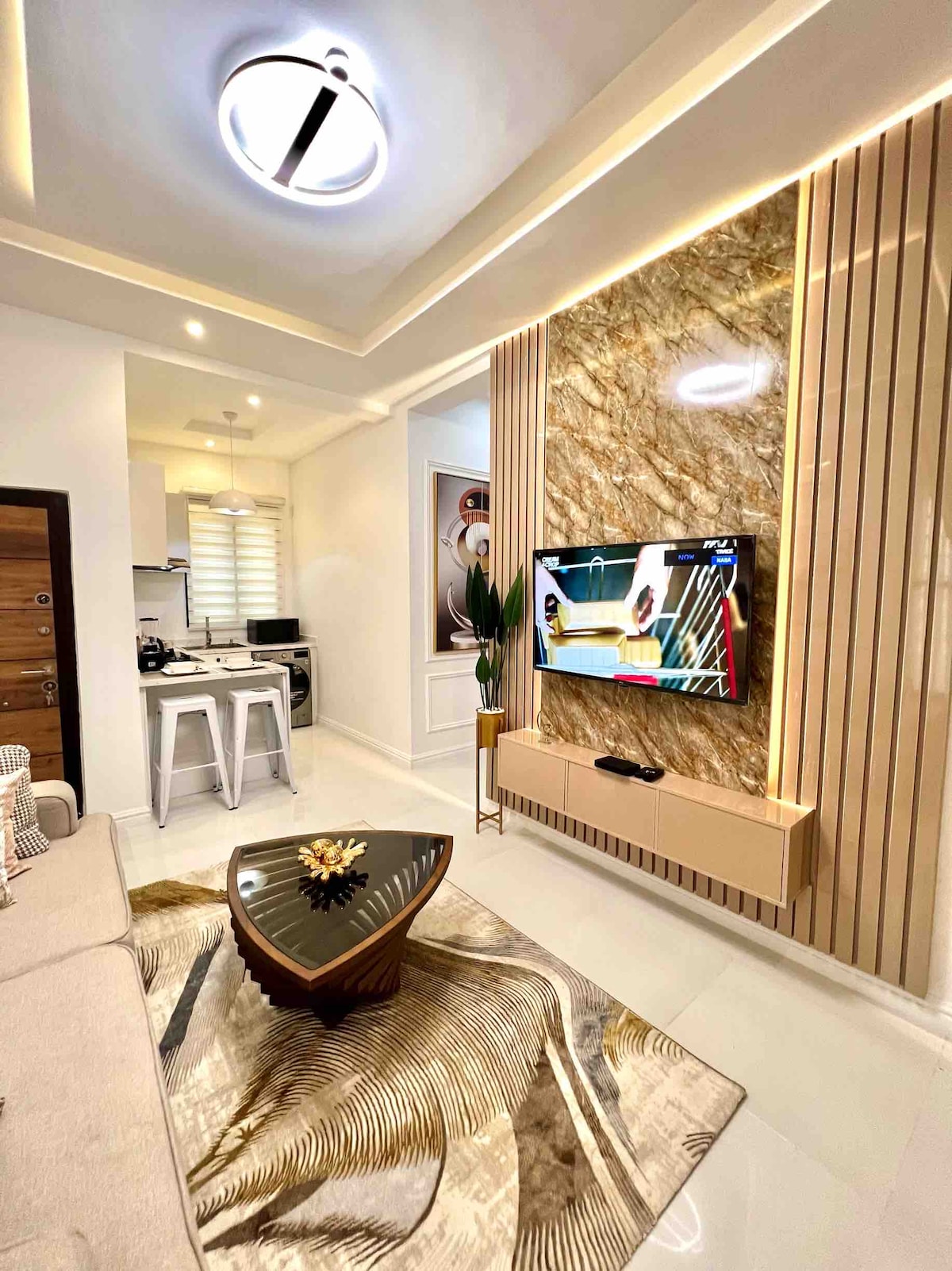 A modern living area is displayed with a plush sofa facing a flat-screen TV mounted on a stylish wall. A decorative coffee table sits atop a patterned rug, and a light fixture illuminates the space. The open layout leads to a kitchenette, enhancing a sense of connectivity.