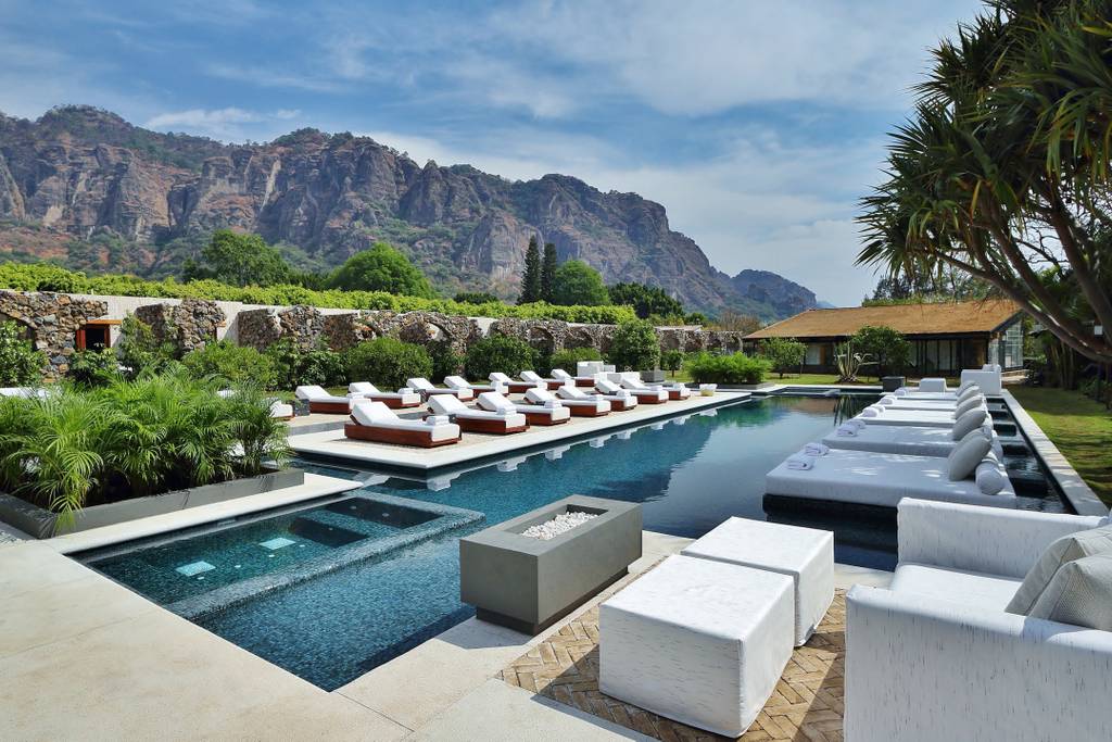 An outdoor area showcases a heated pool surrounded by comfortable chaises. Lush greenery and tall mountains create a serene backdrop. The pool area features both a shallow and deep section, with a sleek design reflecting the clear sky above.