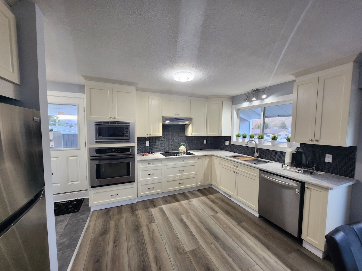 A modern kitchen features white cabinetry and stainless steel appliances, including an oven and dishwasher. Natural light floods the space through a window above the sink, accompanied by minimalist decor of potted plants. The flooring is composed of light-colored wood planks, enhancing the bright environment.