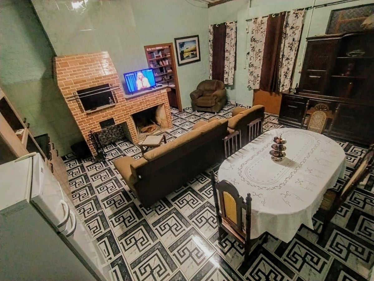 A spacious living area is presented with a large dining table set for meals, comfortable seating arrangements, and a television mounted on the wall. A decorative fireplace is positioned within a brick frame, and large windows are adorned with patterned curtains, allowing natural light to filter through.