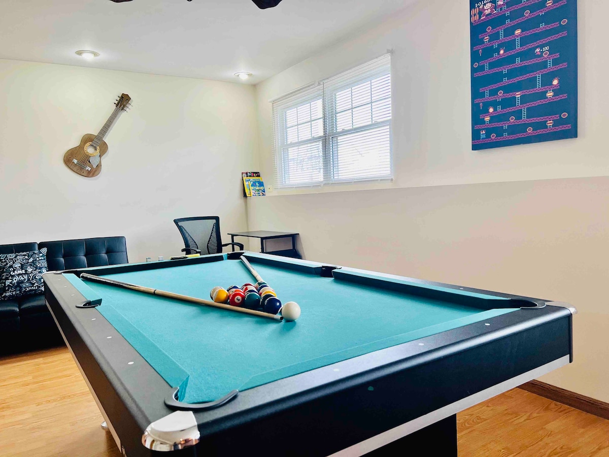 A game room features a billiards table with colorful balls arranged for play, accompanied by a black leather sofa and a rolling chair. Natural light enters through two windows, highlighting a guitar mounted on the wall and a colorful art piece nearby.