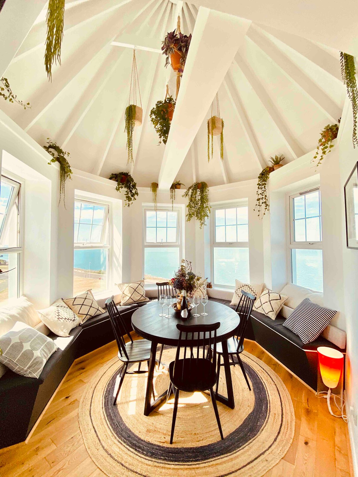 A round dining area features a central table surrounded by black chairs. Bay windows are adorned with plants and decorative cushions, providing ample natural light. A patterned area rug adds warmth to the space, while light wood flooring complements the modern design.
