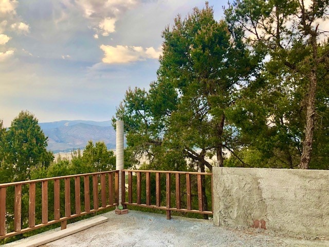 A spacious outdoor deck is surrounded by tall trees, offering views of distant mountains under a partly cloudy sky. The wooden railing enhances the natural setting, while a stone wall provides structure. A chimney is visible, adding to the rustic charm of the area.