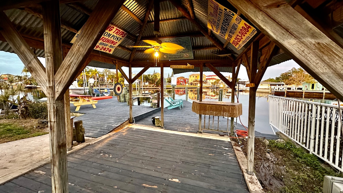 A spacious tiki hut with a weathered wooden structure is shown, featuring a light fixture and open sides for ventilation. Signs are hung from the rafters, while a wooden counter provides space for seating or preparation. The surrounding waterfront view includes docks and grassy areas.