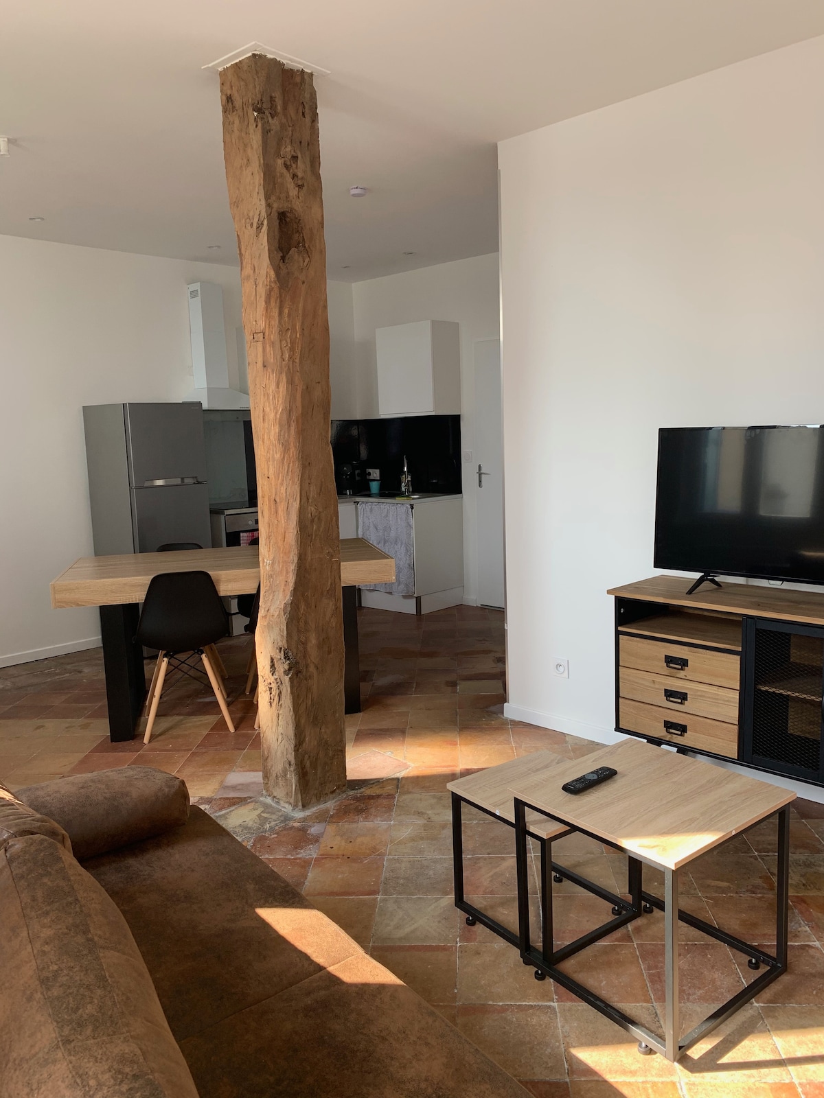 An open living space is highlighted by a central wooden beam, with a wooden dining table and modern black chairs visible. A kitchen area features sleek cabinetry, while a television is positioned on a media console, complemented by varying table heights for functionality.