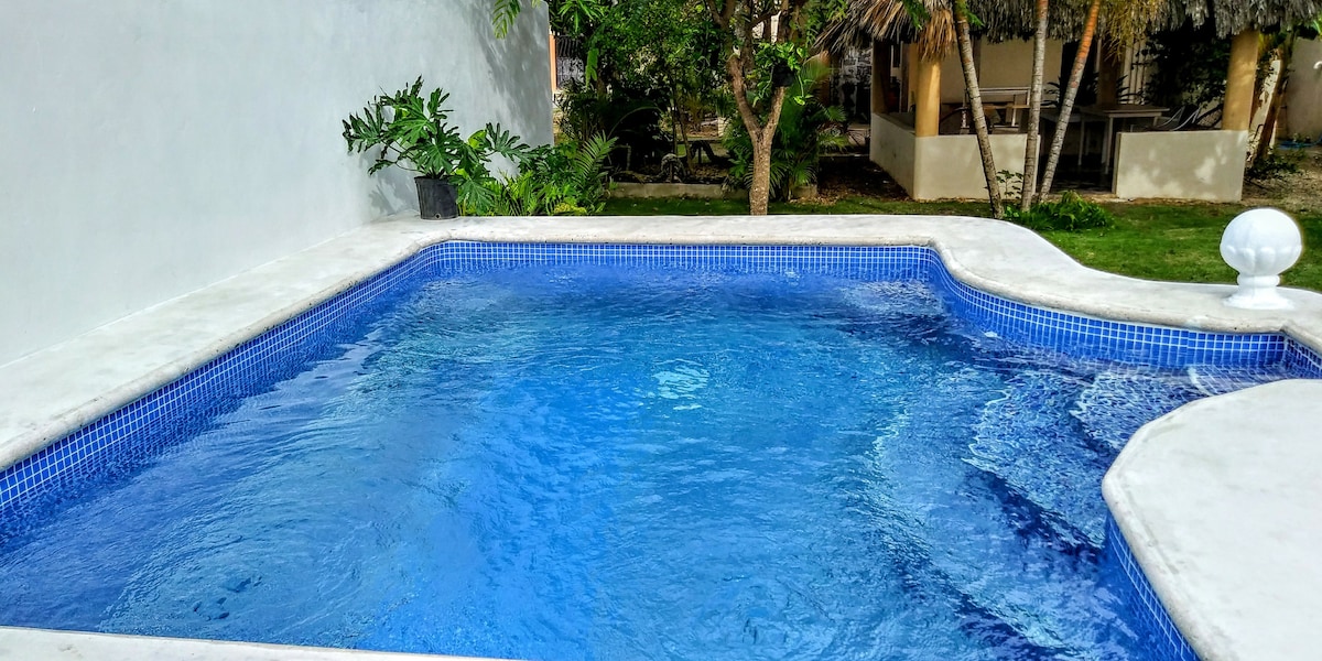 A private pool features clear blue water, surrounded by a smooth concrete deck. Lush greenery is visible nearby, adding to the serene environment. A shaded area can be seen in the background, complementing the outdoor space.