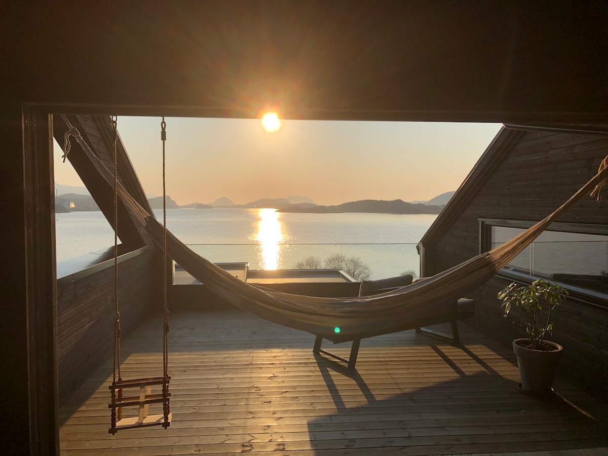 A shaded terrace offers a view of the water at sunset, with sunlight reflecting on the surface. A suspended hammock is visible alongside a small table and plant, creating a comfortable spot for relaxation.