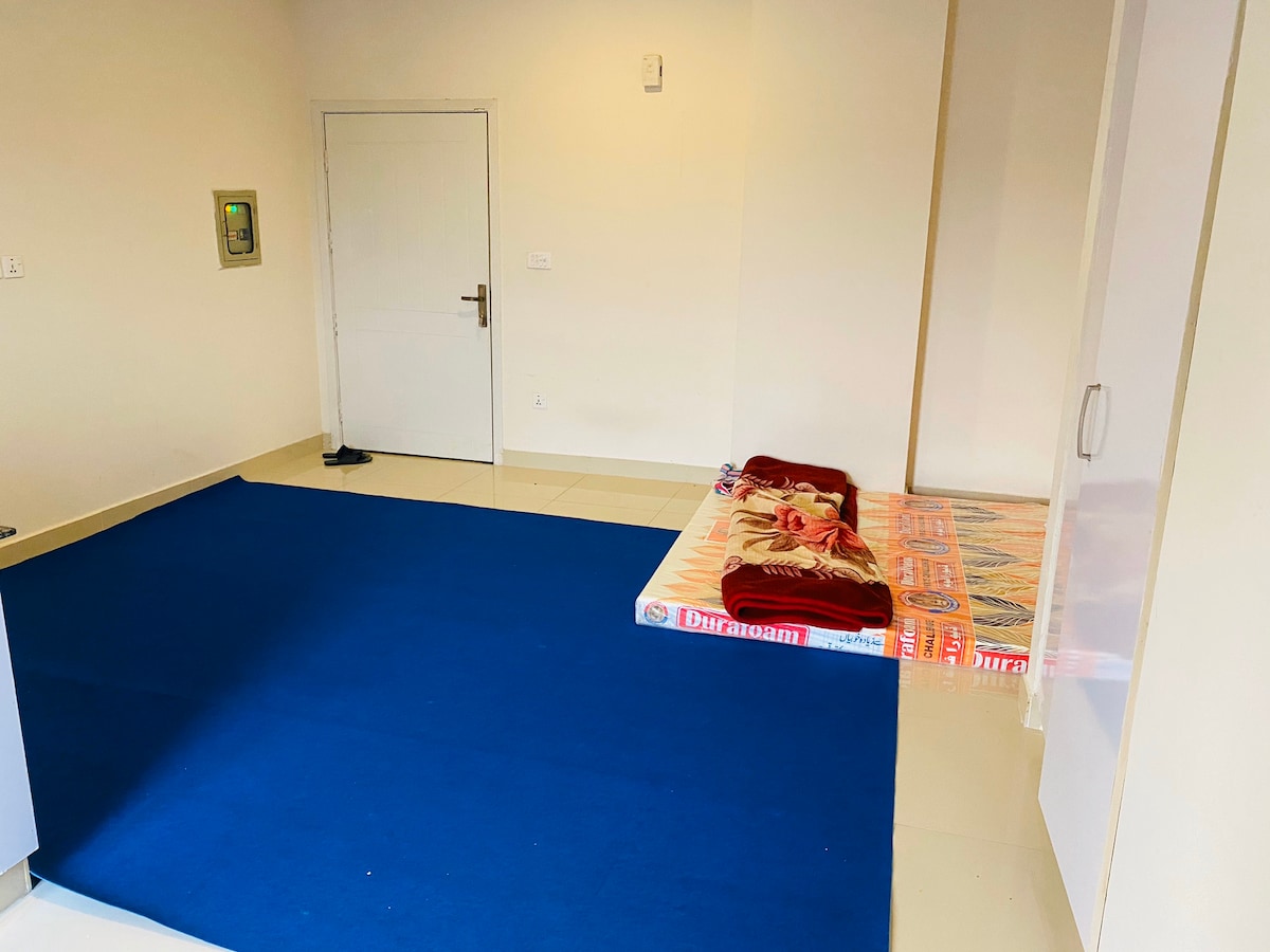 An empty room is presented, featuring a blue carpet that covers part of the floor. A mattress is placed against one wall, adorned with a decorative cover. A closed white door is visible, along with a light-colored cabinet occupying one corner of the room.