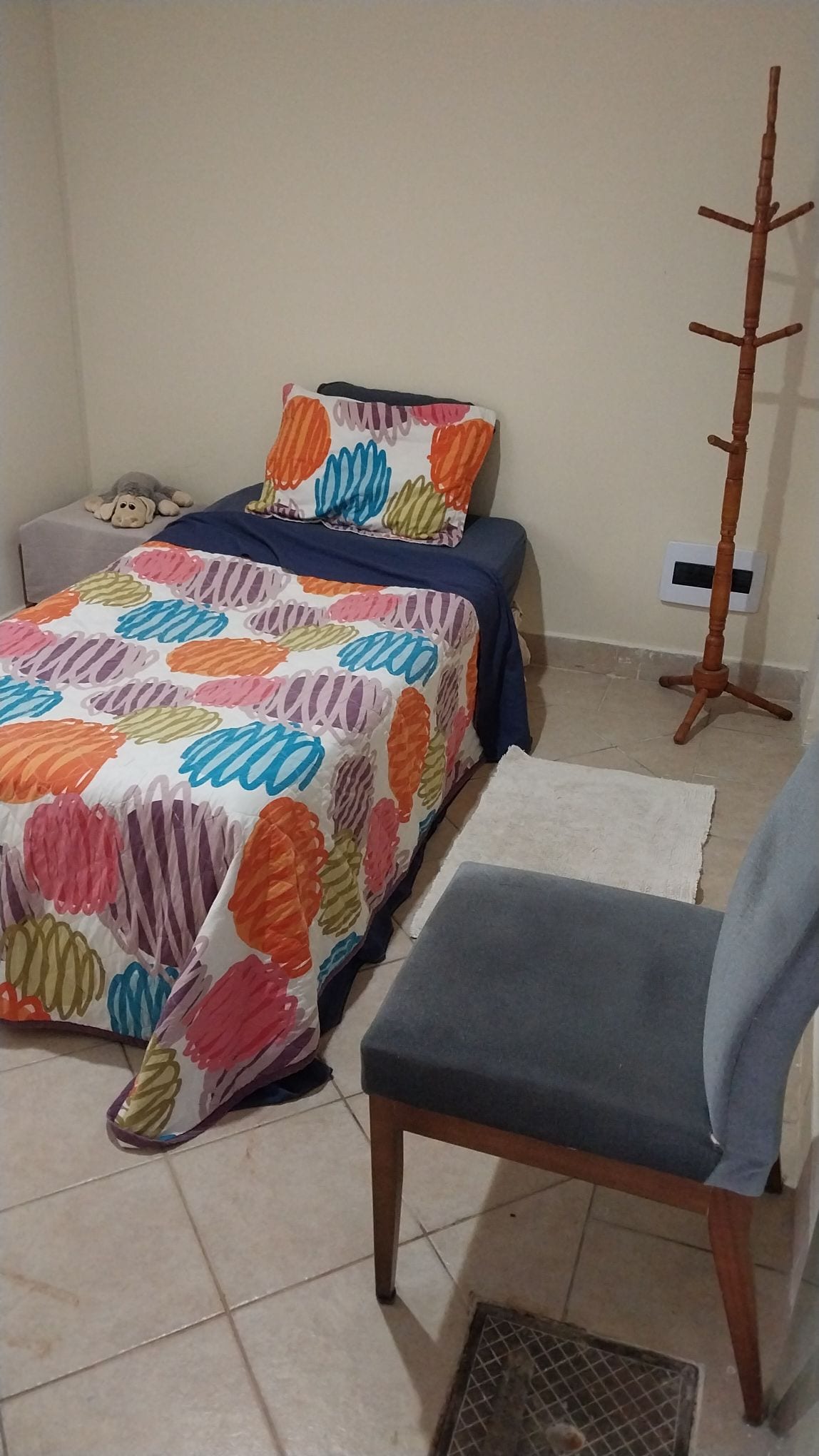 A compact bedroom features a single bed adorned with a colorful patterned blanket and coordinating pillow. A plush toy is placed at the head of the bed. A simple wooden chair sits beside a small nightstand, and decorative wall hooks are mounted nearby.