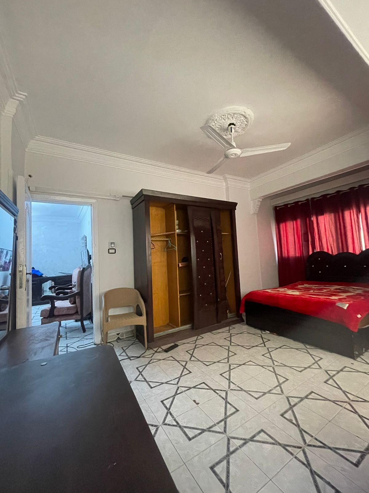 The room features a spacious bed with red bedding and two large windows dressed in deep red curtains. A wooden wardrobe stands beside the bed, and a simple chair is positioned near a dark wooden table. A doorway leads to another room in the background.